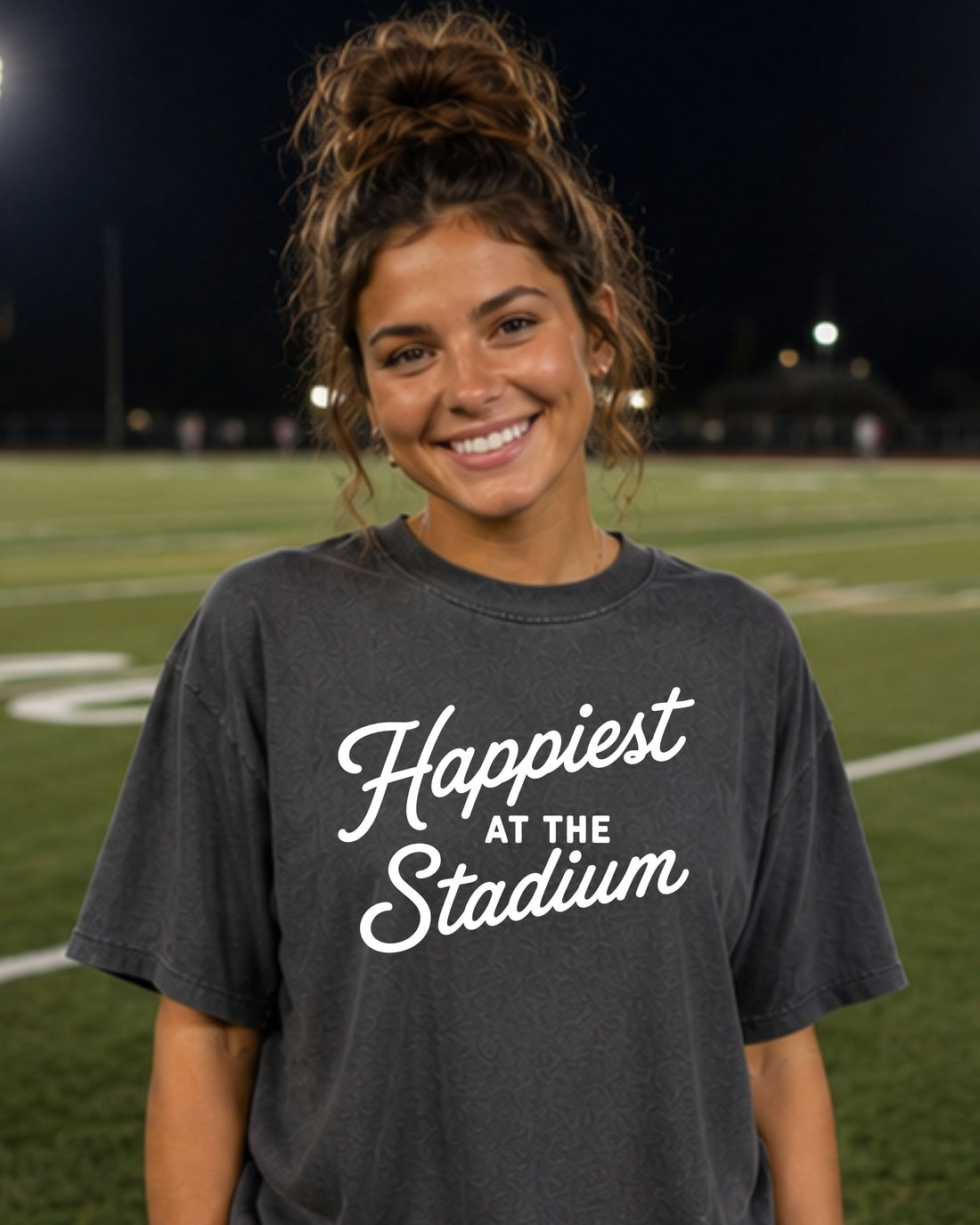 Woman wearing Happiest at the Stadium charcoal Comfort Colors tee on a football field at night
