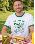 Man wearing a t-shirt with a pickle graphic and text, sitting at a picnic table.