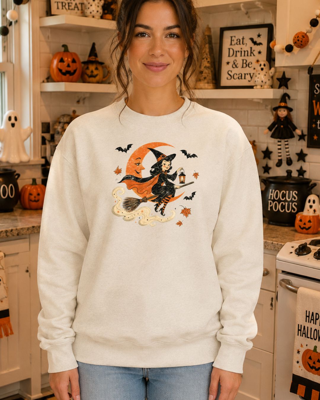 Woman wearing a white crewneck sweatshirt with a vintage-style illustration of a pinup witch riding a broomstick past an orange crescent moon with bats and autumn leaves