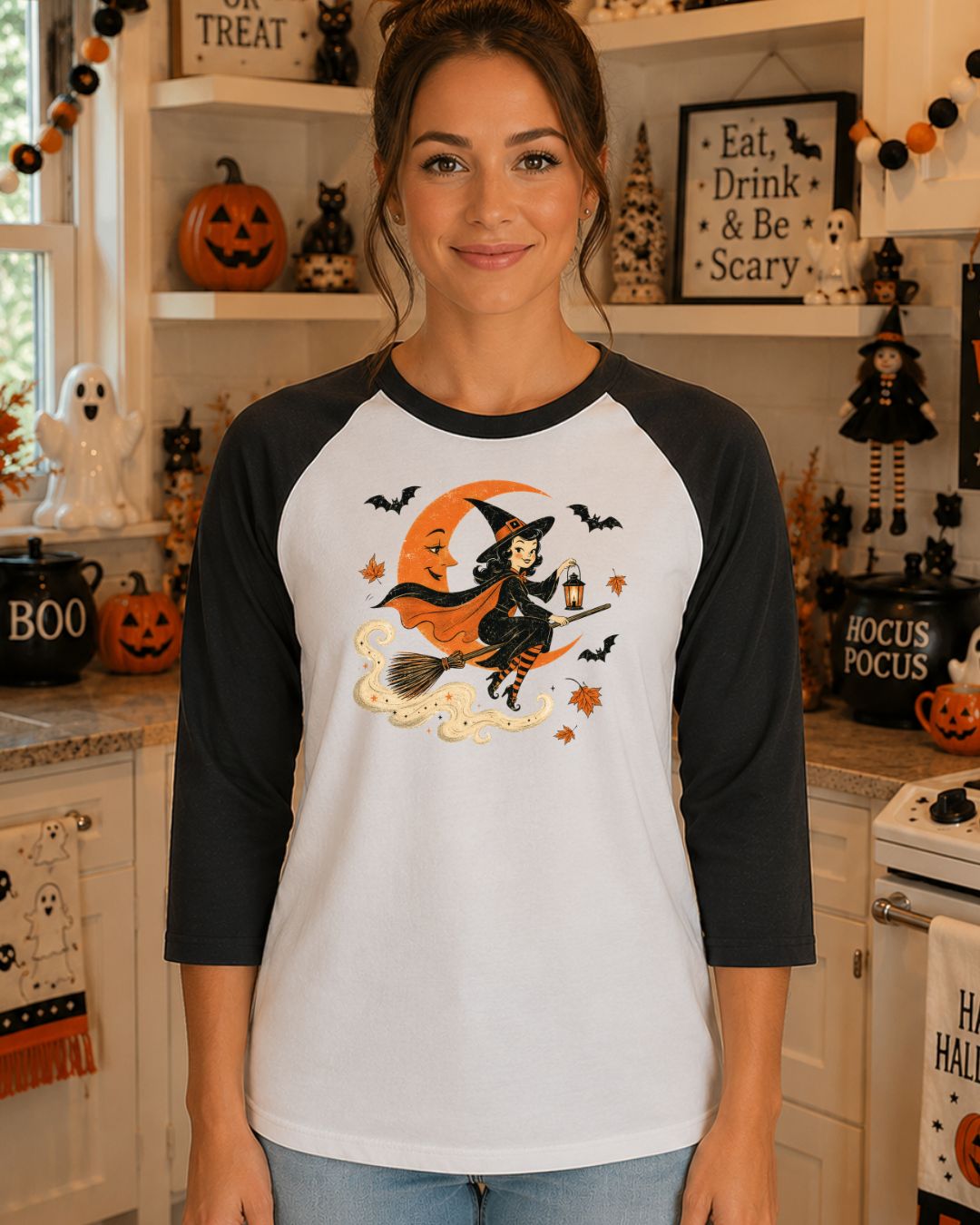 Woman wearing a white and black raglan baseball tee with a vintage-style illustration of a pinup witch riding a broomstick past an orange crescent moon with bats and autumn leaves