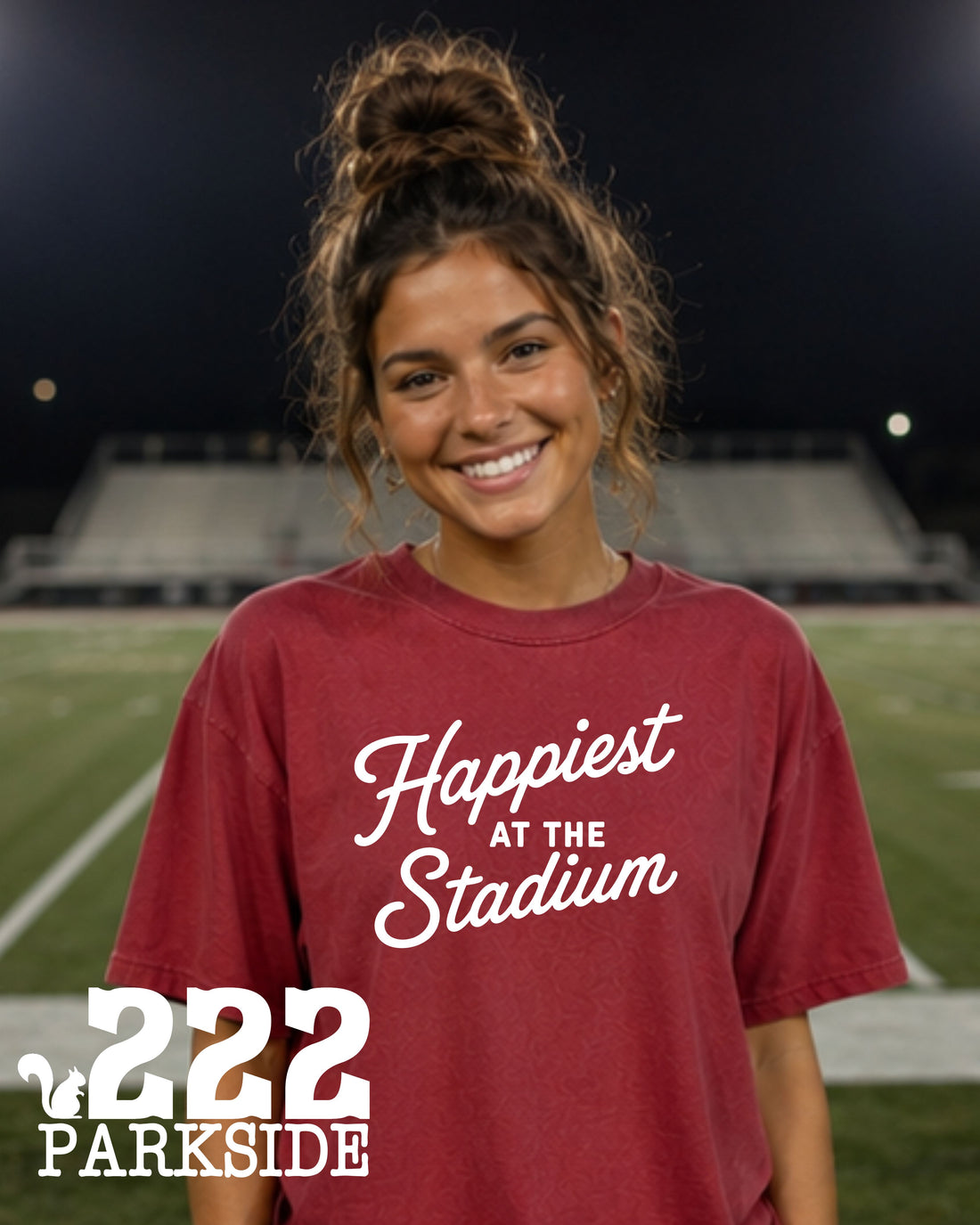 Woman wearing Happiest at the Stadium crimson Comfort Colors tee at a football stadium at night